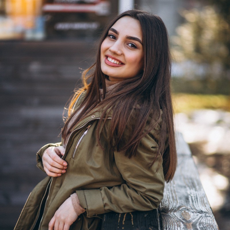  A woman wearing an olive green jacket carries a black bag, standing confidently in a casual setting.