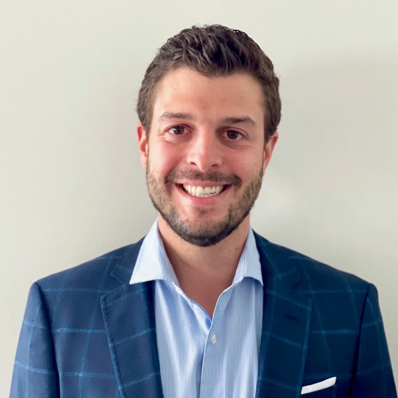 A man wearing a blue suit and white shirt, standing confidently with a neutral expression.
