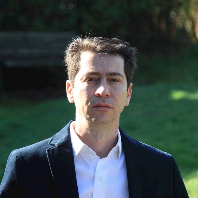 A man in a suit and white shirt stands confidently in a lush green park, surrounded by trees and sunlight.